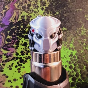 May include: A silver Predator helmet figurine with black dreadlocks and a red dot on the side. The figurine is mounted on a silver and black object, set against a colorful, abstract background.