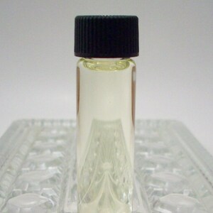 May include: A small clear glass bottle with a black screw-on cap, filled with a light yellow liquid. The bottle is sitting on a clear glass surface.