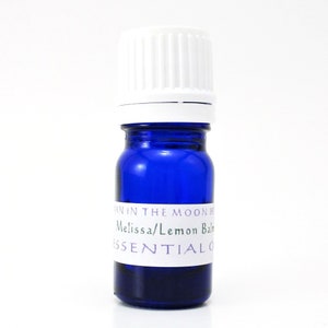 May include: A small blue glass bottle with a white cap. The bottle has a white label that reads "Man in the Moon Herbs Melissa/Lemon Balm Essential Oil".