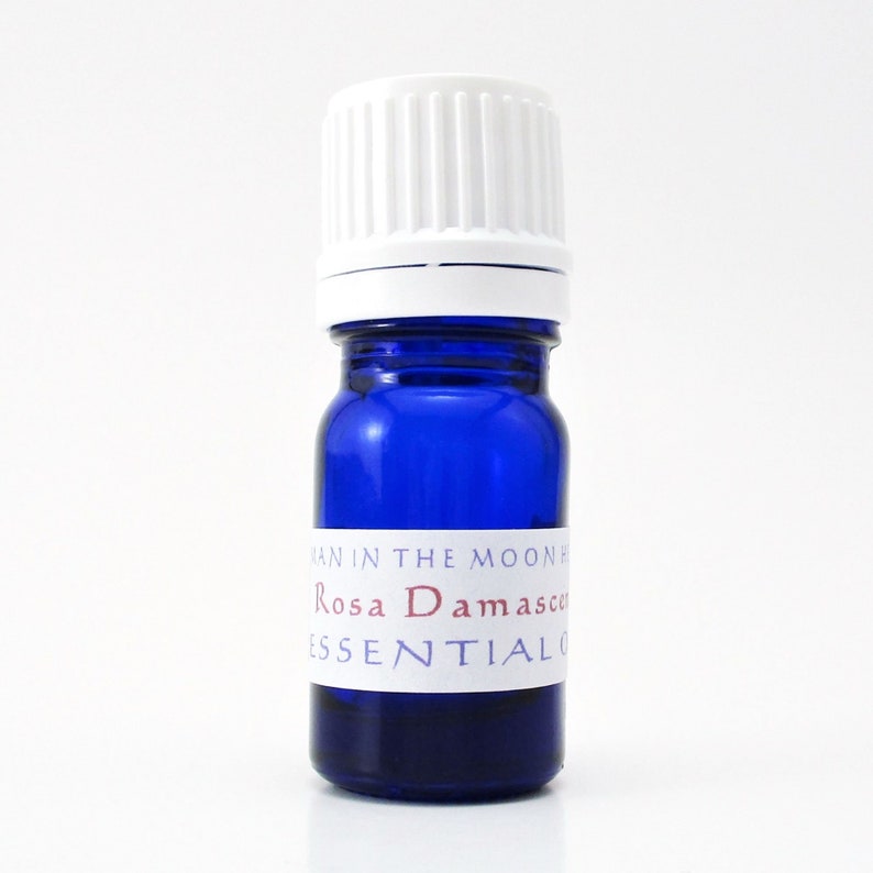 A cobalt blue glass bottle of rose essential oil. The shape of the bottle is squat and the cap is ribbed and white. It is labeled Man in the Moon Herbs Rosa Damascena Essential Oil.