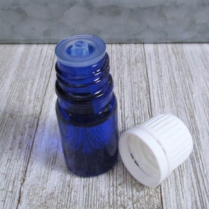 May include: A small blue glass bottle with a white dropper cap and a white screw-on lid. The bottle is sitting on a light wood surface.