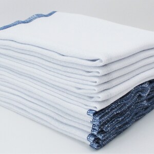 May include: A stack of white cotton dishcloths with blue edging. The cloths are folded and stacked neatly.