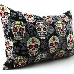 May include: A decorative pillow featuring a repeating pattern of colorful sugar skulls on a black background with gray floral accents. The skulls have intricate designs with red, orange, green, and yellow details. The pillow is rectangular.