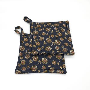 May include: Two square navy blue pot holders with a floral pattern in gold and red. Each pot holder has a small loop for hanging. The pot holders are made of a quilted fabric.