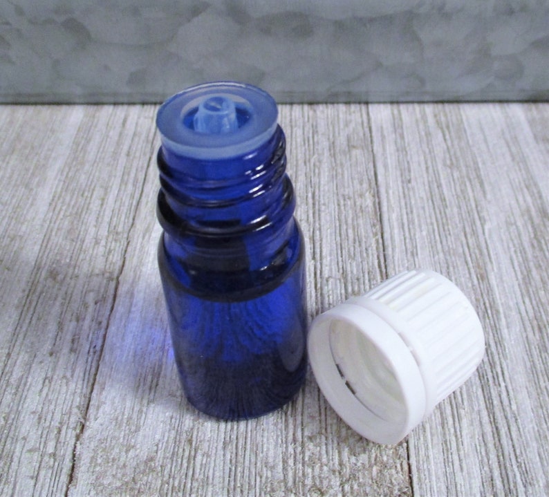 May include: A small blue glass bottle with a white dropper cap and a white screw-on lid. The bottle is sitting on a light wood surface.