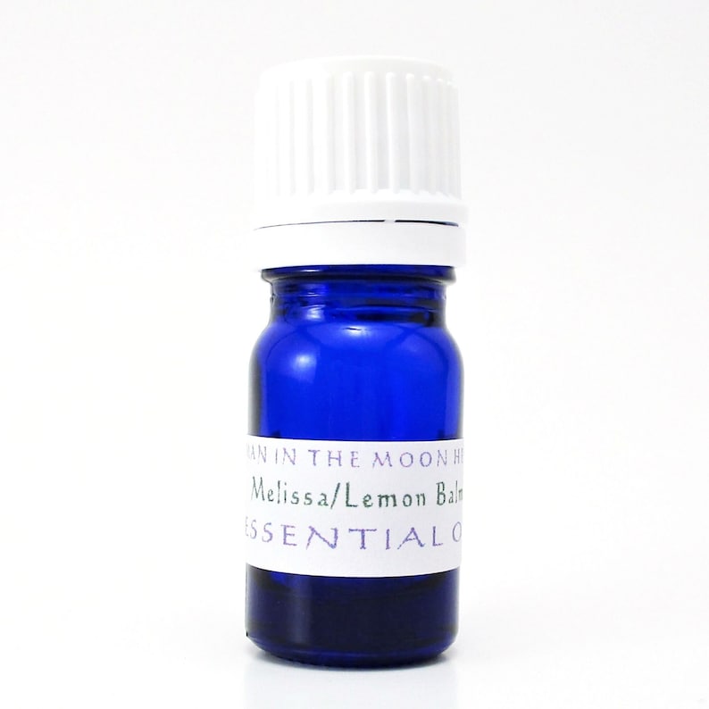 May include: A small blue glass bottle with a white cap. The bottle has a white label that reads "Man in the Moon Herbs Melissa/Lemon Balm Essential Oil".