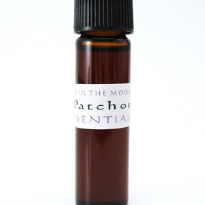 Pode incluir: Um pequeno frasco de vidro castanho com uma tampa preta, etiquetado "Patchouli Essential Oil" em letras brancas com acentos roxos. O r&oacute;tulo tamb&eacute;m diz "In The Moon" em letras menores.