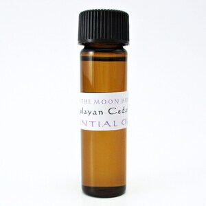 May include: A small amber glass bottle with a black cap, filled with a clear liquid. The bottle has a white label with the text "The Moon Herb Himalayan Cedar Essential Oil".