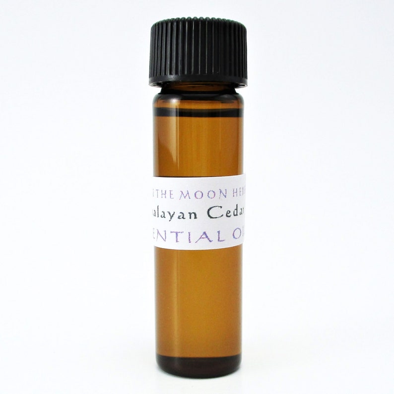 May include: A small amber glass bottle with a black cap, filled with a clear liquid. The bottle has a white label with the text "The Moon Herb Himalayan Cedar Essential Oil".