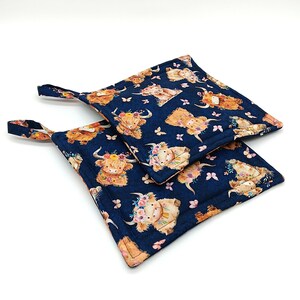 May include: Two square navy blue pot holders with a repeating pattern of Highland cows wearing floral crowns and butterflies. Each pot holder has a fabric loop for hanging.