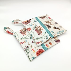 May include: Two square, quilted pot holders with floral and butterfly patterns. The pot holders have a white background with colorful flowers, butterflies, and botanical illustrations. Each pot holder has a teal stripe and a loop for hanging.