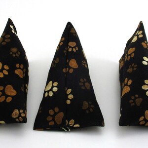 May include: Three black triangular fabric objects with a repeating pattern of tan paw prints. The objects are arranged in a row against a white background. The paw prints vary in size and are evenly spaced.