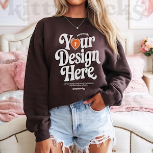 Puede incluir: Una sudadera marrón con el texto "Your Design Here" en una fuente retro con un gráfico de corazón y ojo de cerradura. El texto "kittymocks" aparece arriba y debajo del texto principal.