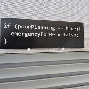 May include: A black rectangular sign with white text that reads "if (poorPlanning == true){ emergencyForMe = false; }". The sign is mounted on a gray textured surface with two white push pins.