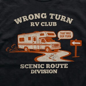 May include: A black t-shirt featuring a graphic of an RV driving on a winding road. The text reads WRONG TURN RV CLUB, SCENIC ROUTE DIVISION, and a speech bubble says THAT WAS OUR TURN. An orange road sign with a white arrow points to the left.