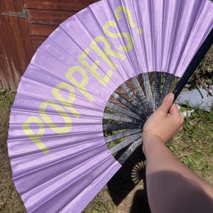 Può includere: Un ventaglio pieghevole color lavanda con la scritta "PER?S" stampata in giallo. Il ventaglio ha un manico nero ed è parzialmente aperto, mostrando il suo design a pieghe. Il ventaglio è tenuto davanti a un capannone di legno e erba verde.