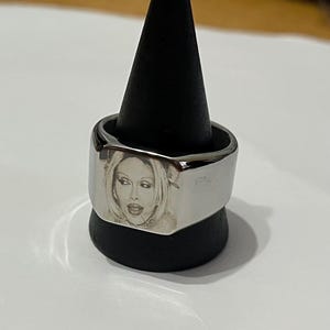 Puede incluir: Un anillo de plata con una cara cuadrada que presenta una imagen grabada en blanco y negro de la cara de una mujer.
