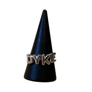 Pode incluir: Um anel prateado com a palavra "DYKE" em letras maiúsculas. O anel é exibido em um suporte de joias preto em forma de cone contra um fundo branco. As letras do anel são em relevo e têm um acabamento brilhante.