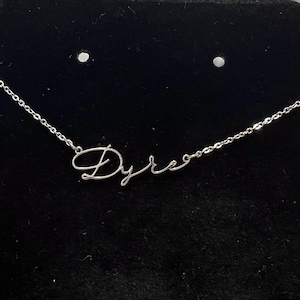 Puede incluir: Un collar de plata con el nombre "Dyre" escrito en cursiva.