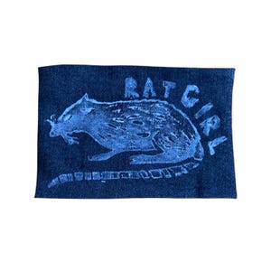 Op de afbeelding: Een rechthoekige, donkerblauwe denim patch met een blauwe illustratie van een rat en de woorden "RAT GIRL" in een handschriftstijl. De rat is in profiel afgebeeld, met zijn staart over de patch.