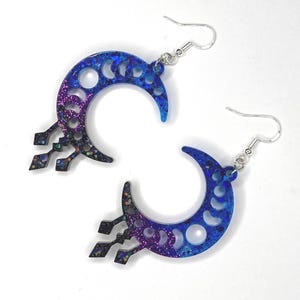 Puede incluir: Un par de pendientes de media luna con un acabado de purpurina azul y morado degradado. Los pendientes presentan un diseño de media luna con un patrón de fase lunar recortado y un dije colgante con un diseño geométrico.