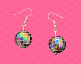 May include: A pair of disco ball earrings. Each earring features a small, spherical disco ball with a mosaic of iridescent squares. The earrings hang from silver-colored fish hook ear wires, set against a bright pink background.