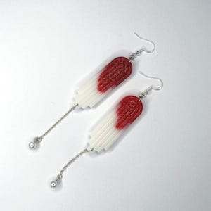 Peut inclure: Une paire de longues boucles d'oreilles pendantes argentées avec des formes géométriques rouges et blanches. Les formes sont en résine et ont une finition pailletée.