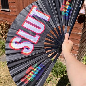 May include: Black folding fan with the word "SLUT" in large, white, 3D-style letters with red and yellow accents. The fan has a rainbow-colored design on the right side. The fan is held open against a wooden background.