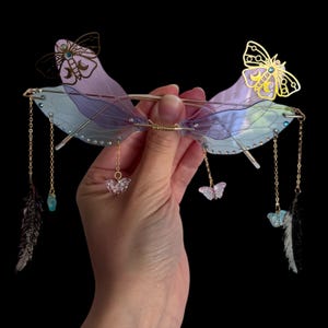 Op de afbeelding: Decoratieve bril met een goudkleurig montuur en iriserende glazen. De bril heeft vlindervleugelvormige accenten in paars en blauw, met gouden vlinderversieringen en bungelende bedels. Kleine kristallen en veermotieven maken het ontwerp compleet.