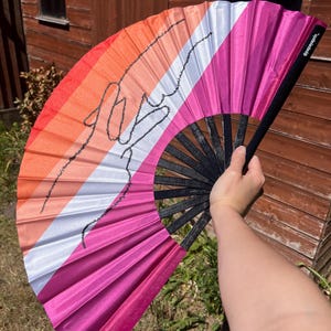 Può includere: Un ventaglio con un design che presenta i colori della bandiera del pride lesbico: arancione, rosa, bianco e magenta. Il ventaglio ha un manico nero e un contorno nero di un disegno. Il testo "@papayaprin_" è stampato sul lato.