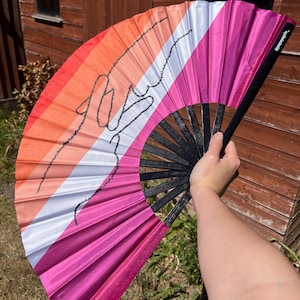 Può includere: Un ventaglio con il design della bandiera del Pride lesbico. Il ventaglio presenta strisce arancioni, rosa, bianche e magenta. Un contorno nero di un disegno è sul ventaglio. Il ventaglio è aperto, mostrando il suo design completo.