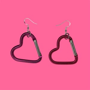 May include: A pair of heart-shaped earrings made from purple metal with silver hooks. The earrings are shaped like a carabiner clip.