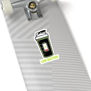 May include: A sticker featuring a black and green shaker bottle design with a low battery indicator. The sticker has the words "LOW BATTERY" in white text on a white background. The sticker is on a skateboard.