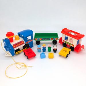 May include: A vintage Fisher-Price Express toy train set featuring a blue locomotive with a yellow pull string, a green freight car, and a red caboose. Includes three small plastic figures, a dog figure, and two small toy cars in red and yellow.