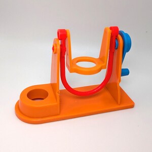 May include: Orange plastic toy swing set with a red rope and blue handle. The swing set has a small orange base with a hole in the center.