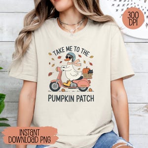 Può includere: T-shirt beige con grafica di un'anatra su uno scooter rosa diretta verso un campo di zucche. L'anatra indossa un casco e una sciarpa. Il testo recita "Take Me to the Pumpkin Patch."
