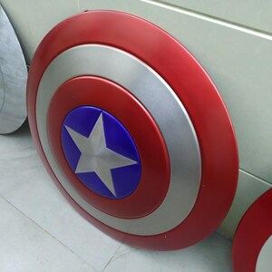 May include: Two red, white, and blue Captain America shields. The shields are round and have a silver star on a blue background in the center. The shields are made of metal and have a shiny finish.