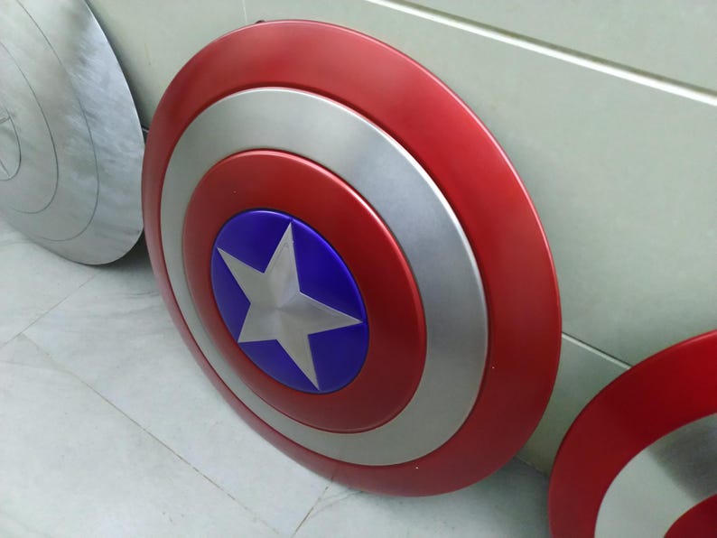 May include: Two red, white, and blue Captain America shields. The shields are round and have a silver star on a blue background in the center. The shields are made of metal and have a shiny finish.
