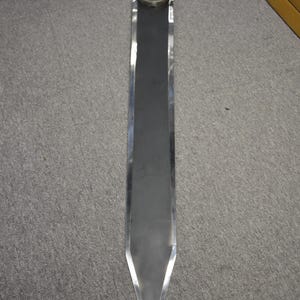 May include: A large, silver-colored sword with a wide, rectangular blade and a pointed tip. The handle is wrapped in white material, and a white fabric is tied to the top. The sword is displayed on a gray surface.