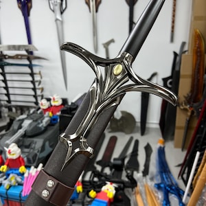 May include: A black and silver sword with a brown leather scabbard. The sword has a decorative hilt with a gold accent. The sword is displayed against a wall with other swords.