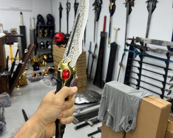 May include: A silver dagger with a gold handle and a red gemstone. The dagger has a textured blade and a pointed tip. The dagger is held in a hand against a background of other swords and shields.