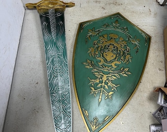 May include: A decorative sword and shield set. The sword features a gold hilt and a blade with intricate teal and silver designs. The shield is teal with gold accents and a central emblem. The set is designed for display.