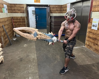 May include: A large, ornate prop weapon with a bone-colored blade and blue accents. The weapon is held by a person wearing a helmet. The helmet is silver with red accents. The person is wearing camouflage shorts and black sneakers.