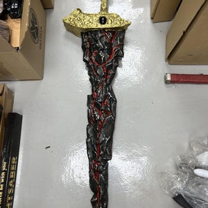 May include: A black and gold sword with a red lava-like design. The sword has a gold hilt and a black blade with red streaks running down the length of the blade.