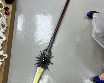 May include: A spear with a long, dark red shaft and a silver and gold blade. The spear features a decorative black sunburst design near the blade and a gray handle. The spear is lying on a gray surface.