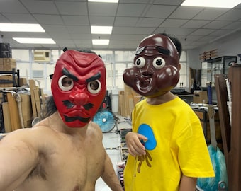 May include: Two people wearing masks. One person is wearing a red mask with a frowning expression and the other person is wearing a brown mask with a surprised expression. The person wearing the brown mask is wearing a yellow shirt with a blue and yellow graphic.
