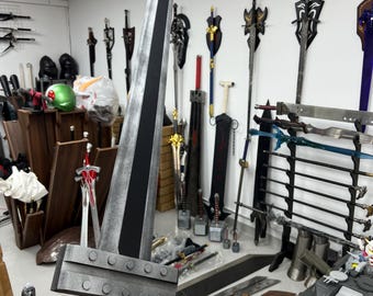 May include: A large, black and silver sword with a white handle. The sword is held by a person's hand. The sword is in a room with many other swords on the wall.