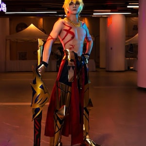 May include: A person in a cosplay costume with a golden blonde wig, gold and red armor, and a large spear. The costume features intricate gold detailing and red accents. The background is a brightly lit indoor space.
