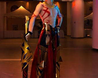 May include: A person in a cosplay costume with a golden blonde wig, gold and red armor, and a large spear. The costume features intricate gold detailing and red accents. The background is a brightly lit indoor space.
