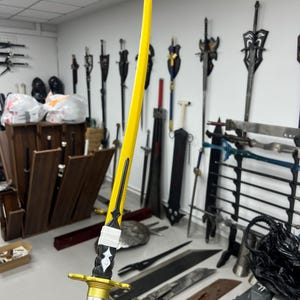 May include: A yellow katana sword with a black and silver handle. The sword is displayed against a wall with other swords.